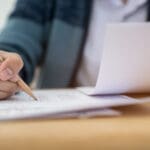 education test concept : hands with blue pen over application form, students taking exams, writing examination room with undergraduate inside. student sitting learning lessons doing final in classroom